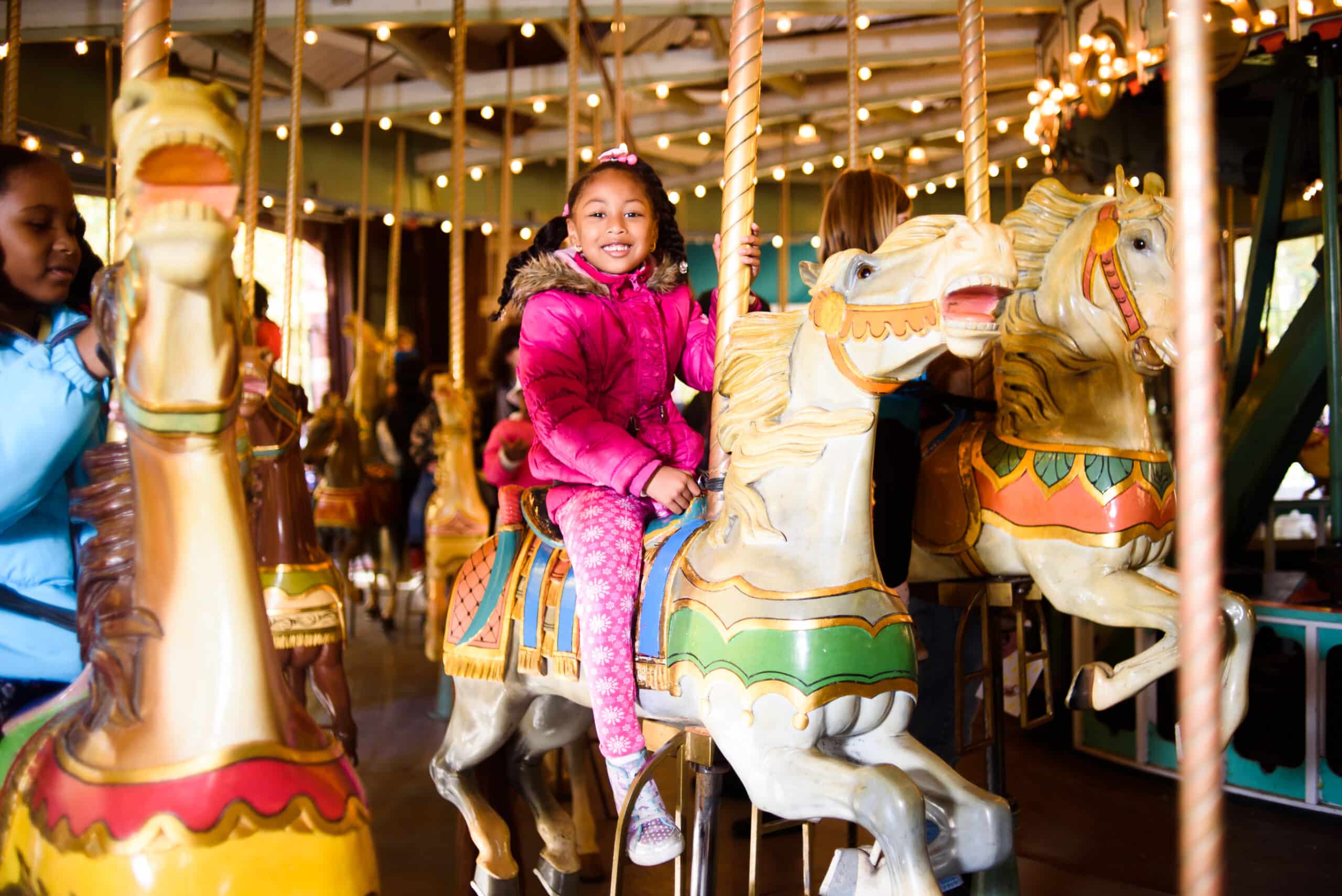 A young girl in a pink coat smiles while riding a horse on the Carousel.