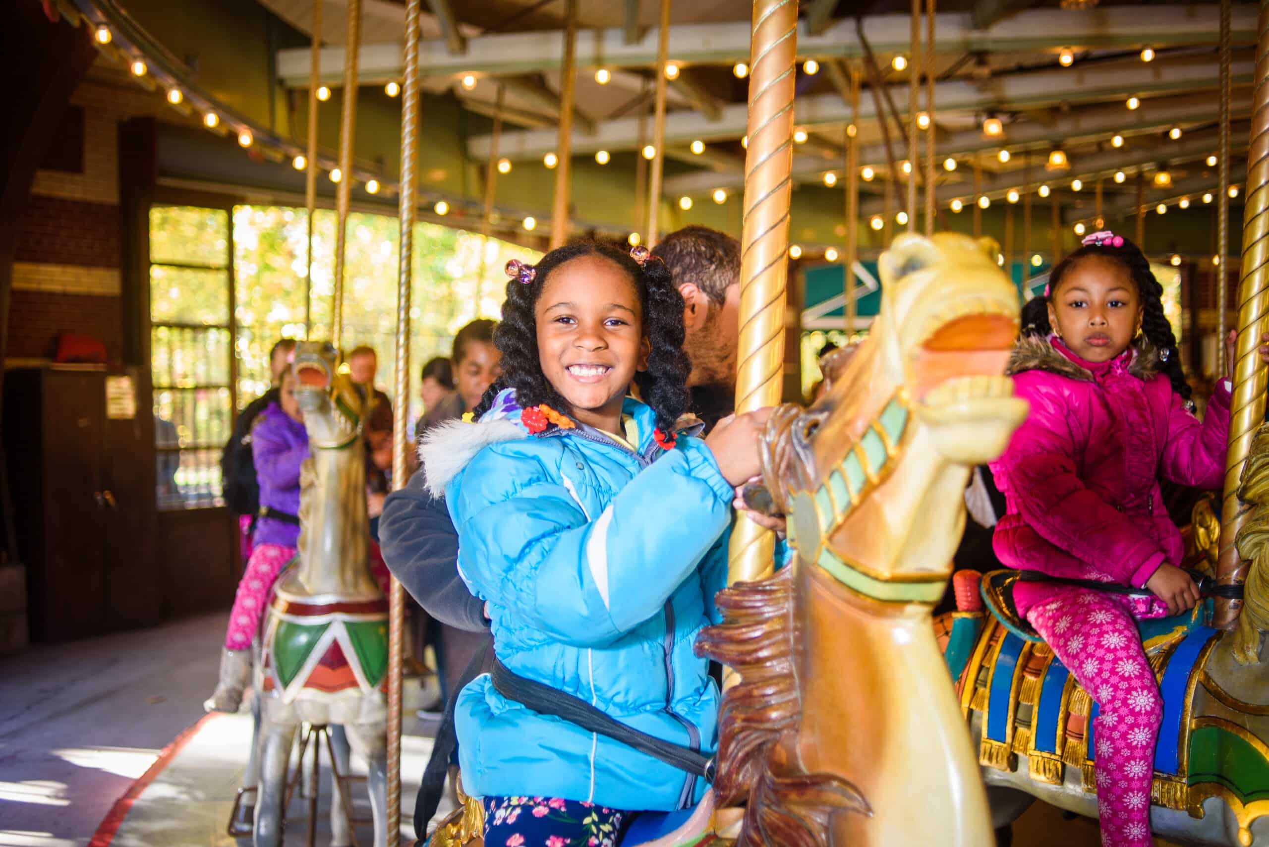 A young girl in a blue coat smiles while riding a horse on the Carousel.