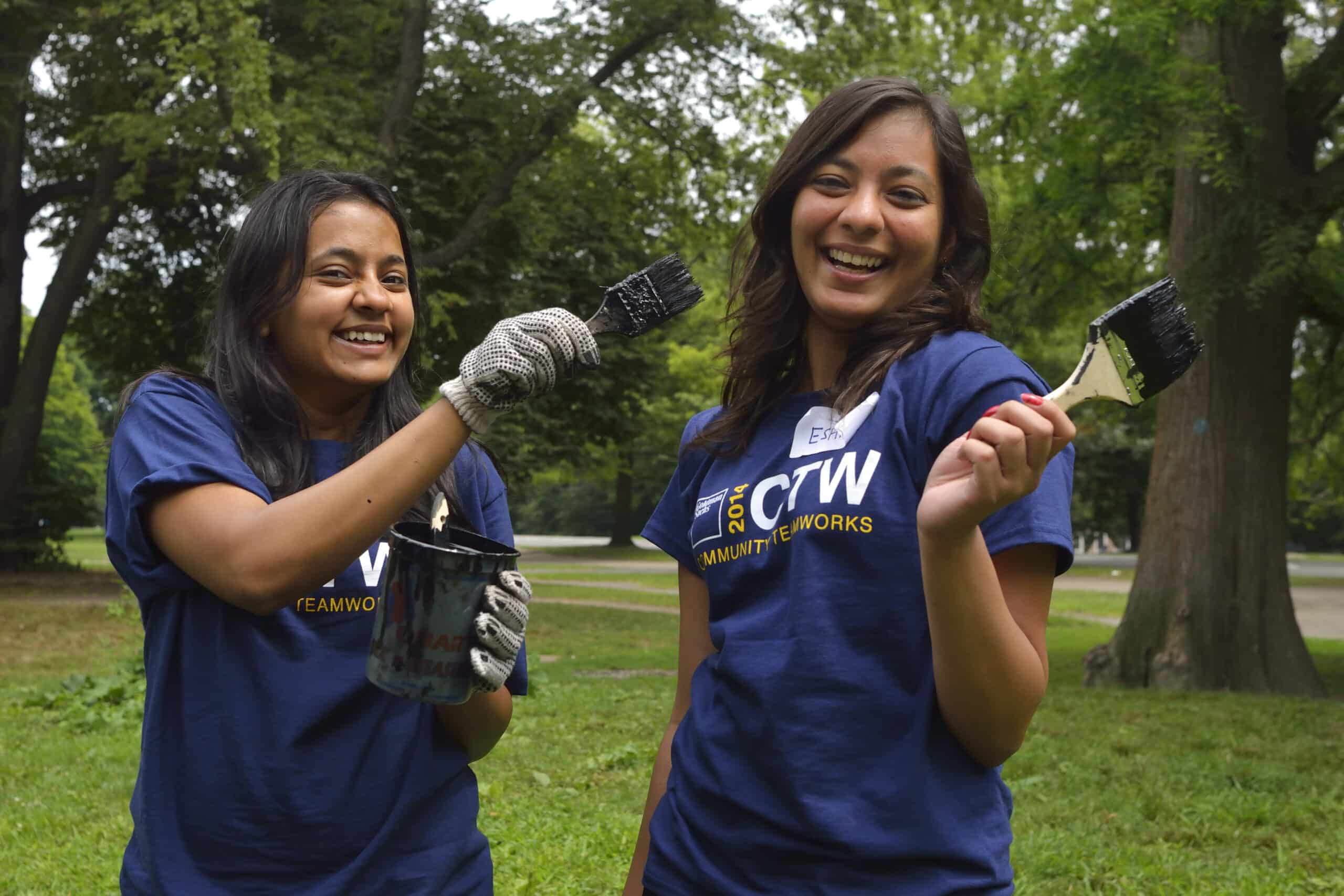 Two women smile holding paint cans and paint brushes in a grassy area of the park.