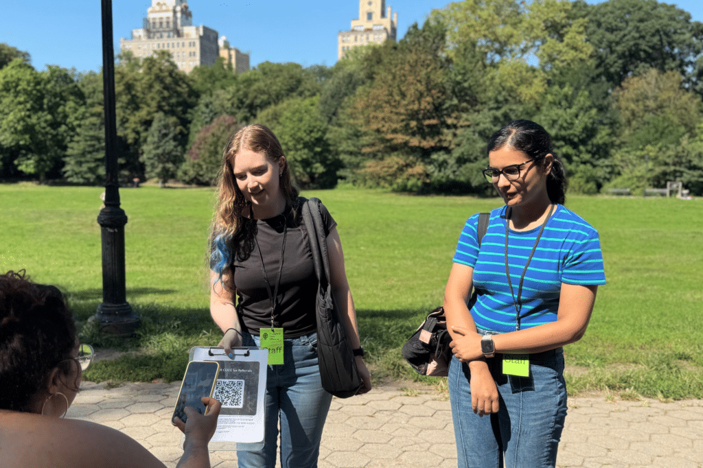 two women with staff badges stand in the park and hold out a clipboard for someone to scan a QR code
